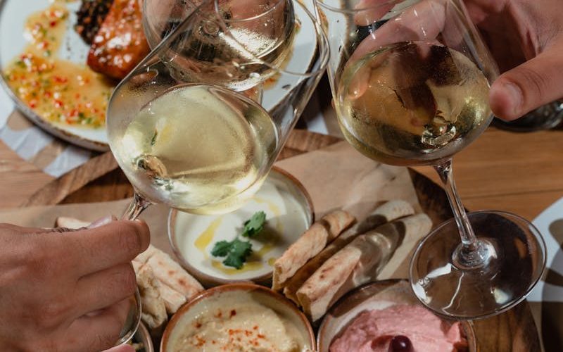 Three adults toast with wine over a table of Mediterranean appetizers including hummus and tzatziki.