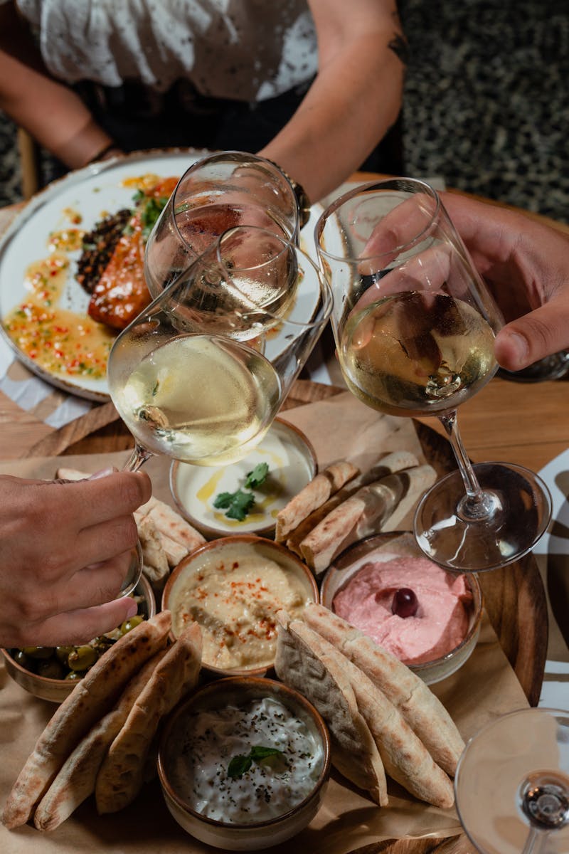 Three adults toast with wine over a table of Mediterranean appetizers including hummus and tzatziki.