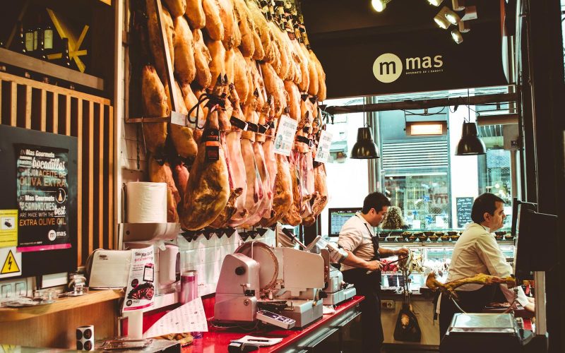 A bustling Spanish ham shop with hanging cured meats and two men serving customers.
